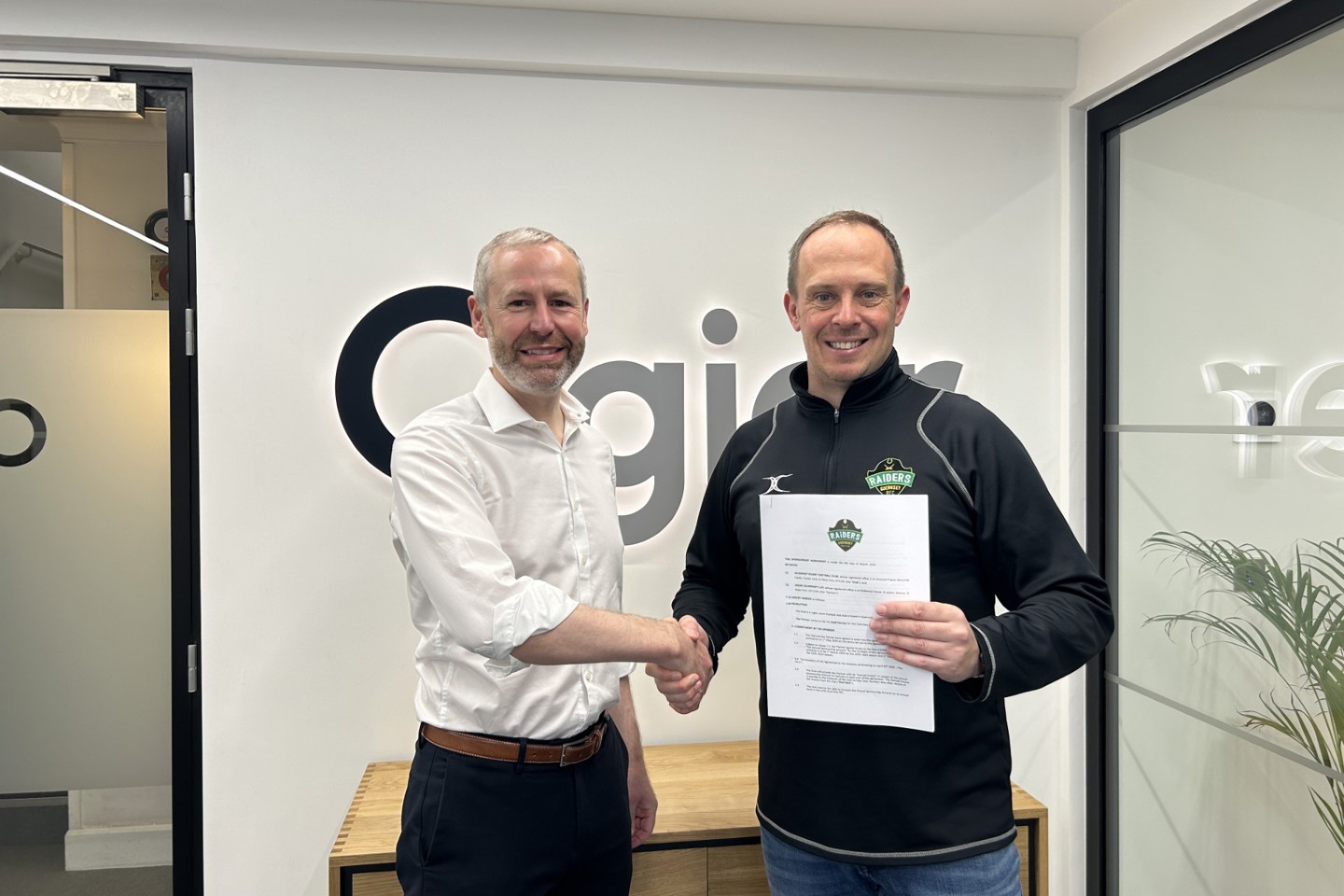 Two people shake hands in front of Ogier logo as Guernsey Raiders sponsorship agreement is signed