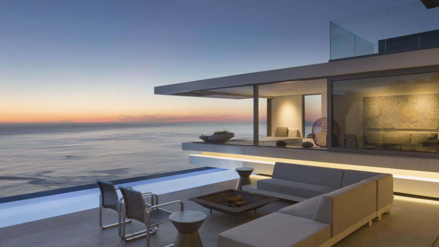 Illuminated modern luxury home with exterior patio, sofa, lap pool, and ocean view at dusk