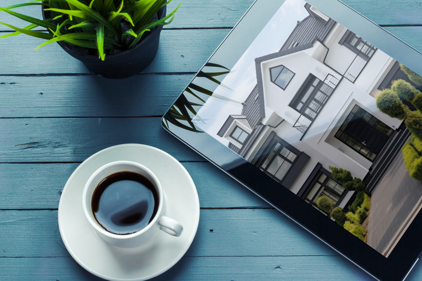 black coffee, plant and an iPad with photo of a house on the screen on top of a blue wooden table
